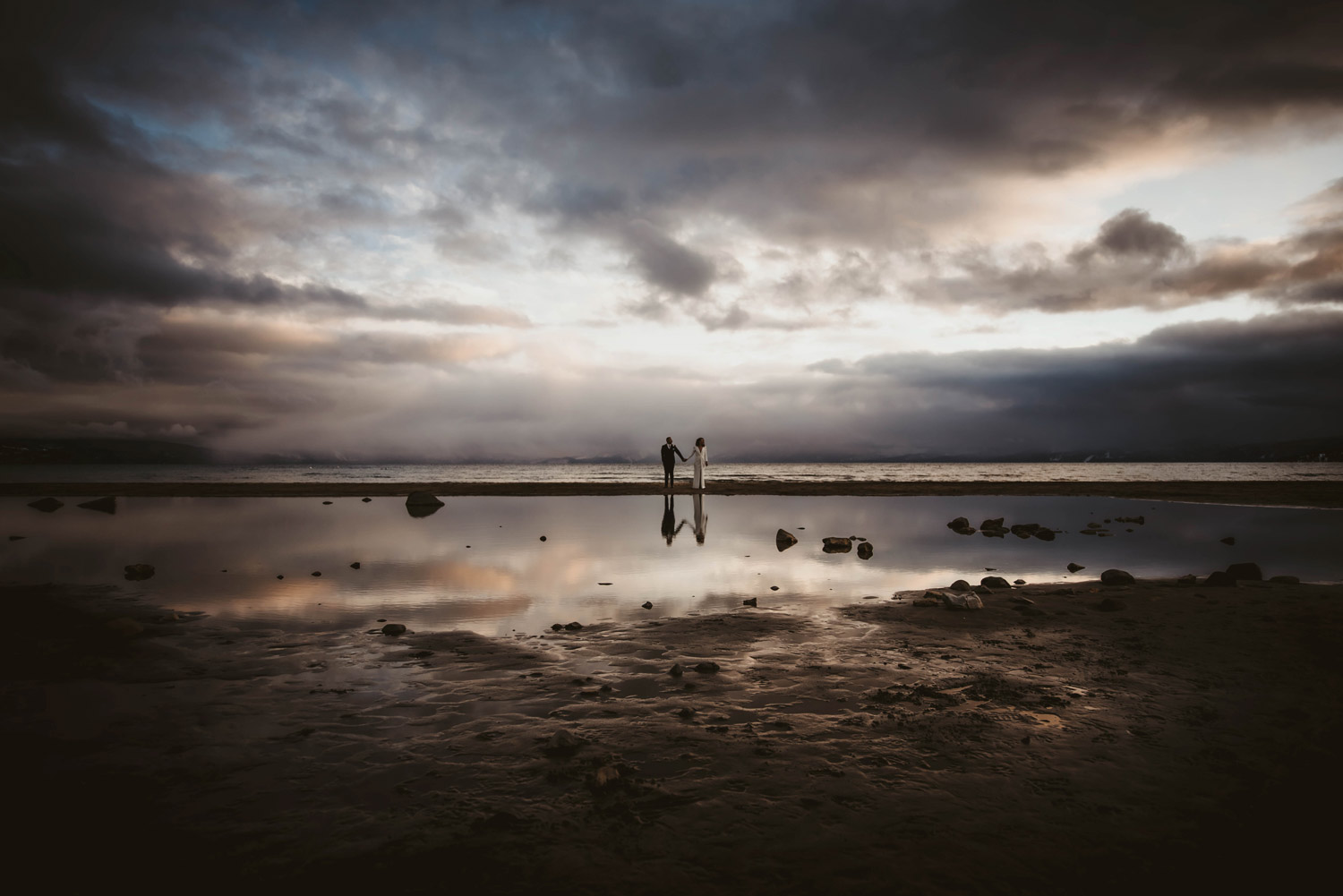moody day on lake tahoe with bride and groom holding hands on shore