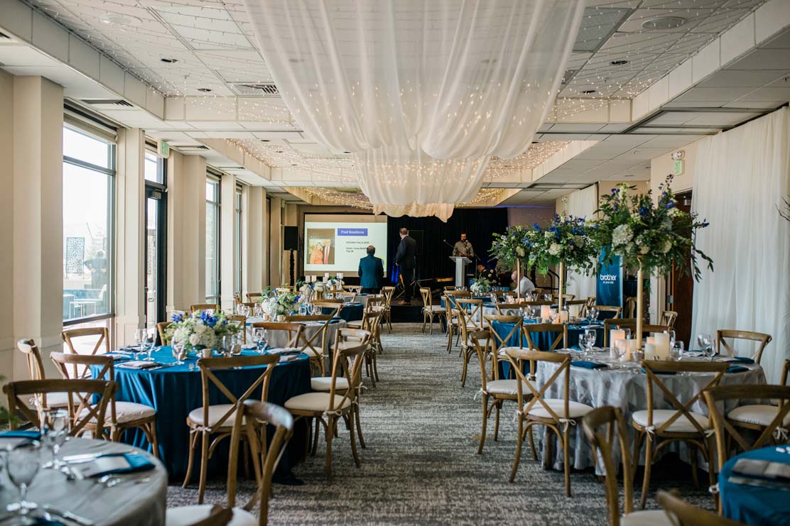 Elegant dining area with royal blue accents, draped fabric and lighting, and a stage with a screen for a slideshow