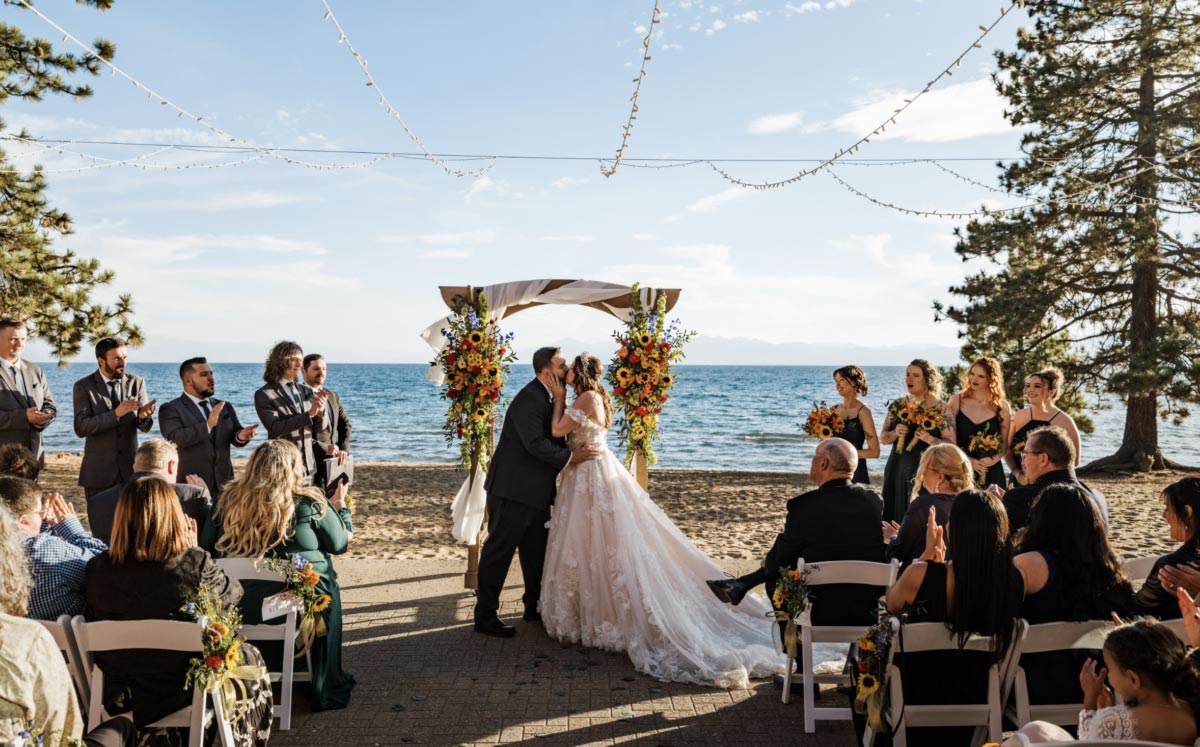 Bride and groom sharing a kiss under a floral arch with guests cheering and clapping set at Lake Tahoe