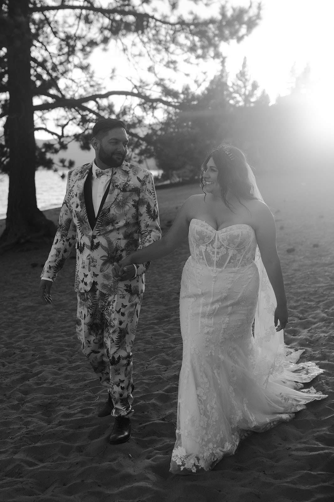 Groom in a bold, patterned suit and bride in a lace dress looking at each other and holding hands