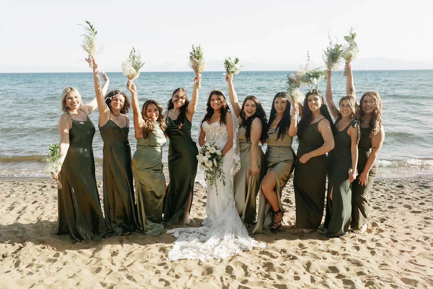 Bride and her happy bridesmaids wearing shades of green, holding floral bouquets in the air with Lake Tahoe in the background