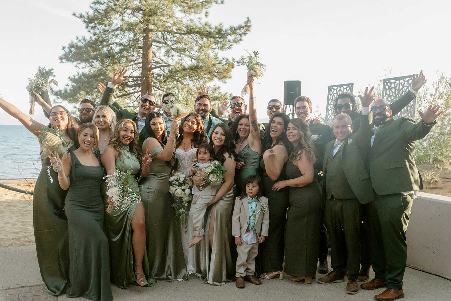 Excited bridal party in green posing together for a group shot in front of Lake Tahoe