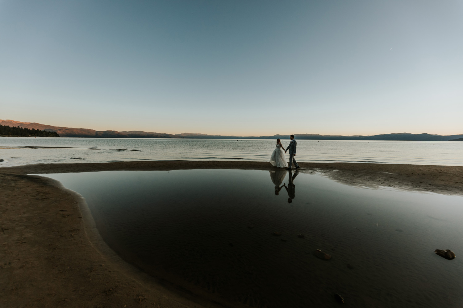 Bride and groom holding hands and walking along the shore of Lake Tahoe at sunset, with dark lighting and their silhouettes reflected on the water