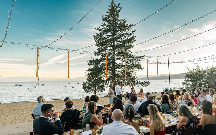 Man giving a speech at an outdoor wedding reception on a terrace filled with guests, with string lights overhead and a lake view in the background.