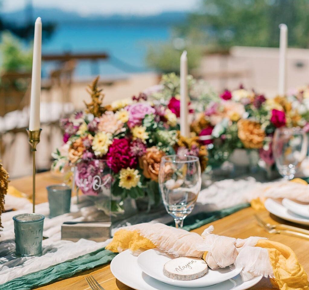 Colorful table settings with bright yellow, pink, and orange decor, white elegant candles, and a blurred view of Lake Tahoe in the background