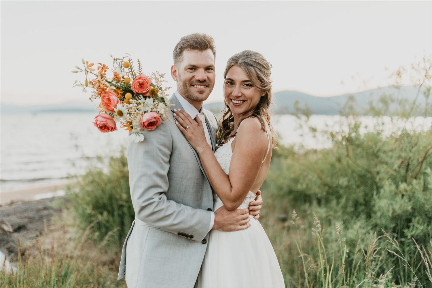 Elated bride holding a coral bouquet and hugging the groom in front of Lake Tahoe