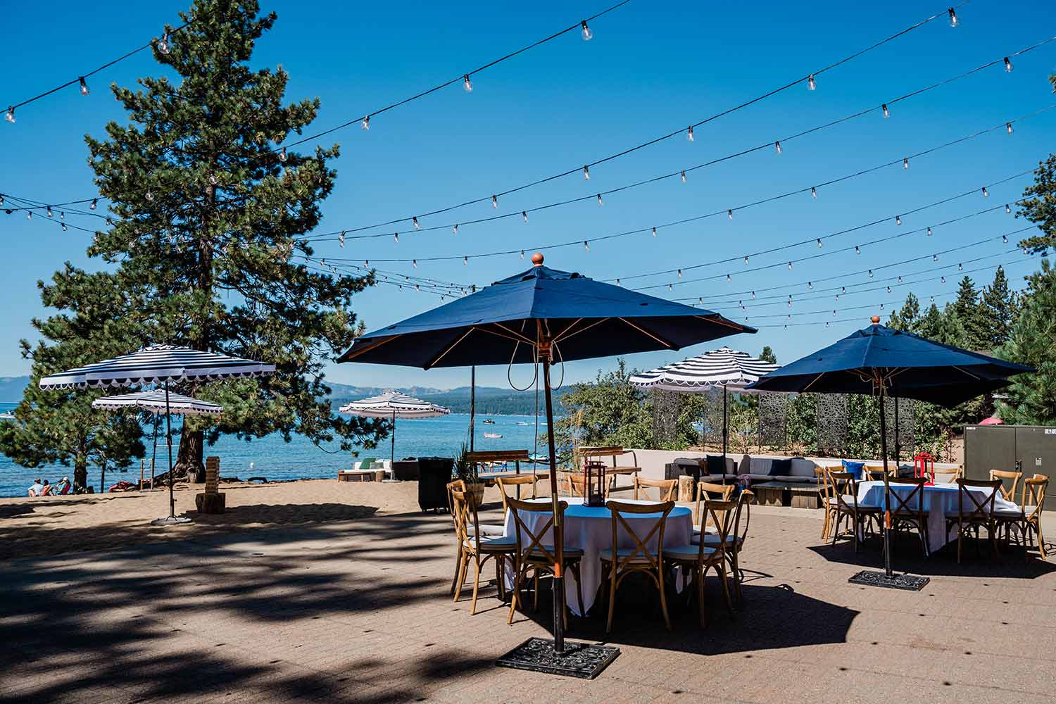 Outdoor terrace with lake view featuring wooden chairs, white linens, a cozy sitting area, and market lights strung overhead